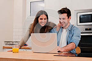 Young caucasian couple browsing on internet at home kitchen using a laptop. Happy husband and cheerful wife planning the