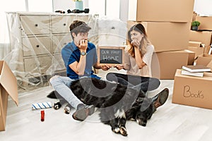 Young caucasian couple with dog holding our first home blackboard at new house looking stressed and nervous with hands on mouth