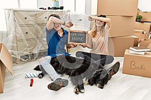 Young caucasian couple with dog holding our first home blackboard at new house smiling cheerful playing peek a boo with hands