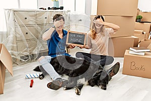 Young caucasian couple with dog holding our first home blackboard at new house smiling and laughing with hand on face covering