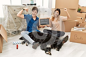 Young caucasian couple with dog holding our first home blackboard at new house strong person showing arm muscle, confident and