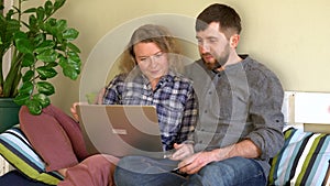 Young Caucasian couple doing shoppings online on the couch at home with laptop, looking at each other and smiling