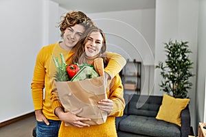 Young caucasian couple holding groceries paper bag hugging each other at home