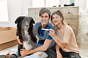 Young caucasian couple holding pregnancy test sitting on the floor with dog at new home