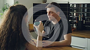 Young caucasian couple sitting at table in modern kitchen dicussing something, curly woman is sitting back to camera