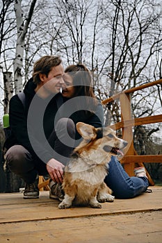 A young Caucasian couple sitting on a wooden bridge in the park, smiling at each other with their dog welsh corgi