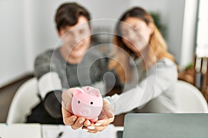 Young caucasian couple smiling happy holding piggy bank sitting on the table at home