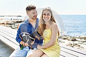 Young caucasian couple smiling happy sitting on the bench with dog at the beach