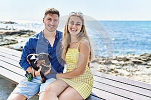 Young caucasian couple smiling happy sitting on the bench with dog at the beach