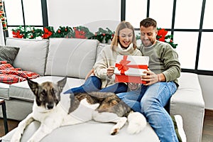 Young caucasian couple smiling happy unboxing gift sitting on the sofa with dog by christmas tree at home