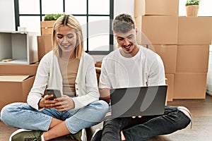 Young caucasian couple smiling happy using laptop and smartphone sitting at new home