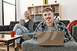 Young caucasian couple smiling happy using laptop and smartphone sitting on the sofa at home