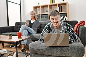 Young caucasian couple smiling happy using laptop and smartphone sitting on the sofa at home