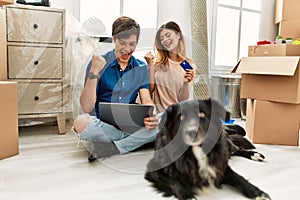 Young caucasian couple using laptop and credit card sitting on the floor with dog at new home