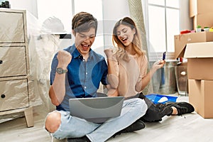 Young caucasian couple using laptop and credit card sitting on the floor at new home