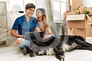 Young caucasian couple using laptop sitting on the floor with dog at new home