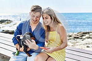 Young caucasian couple using smartphone sitting on the bench with dog at the beach