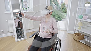 Young Caucasian crippled woman in wheelchair gaming online using motion controllers and VR headset. High angle view of