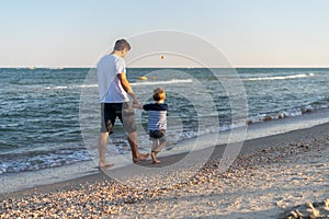 Young Caucasian dad with little son walk warm summer day along the sea coast. Summer family vacation concept. Friendship