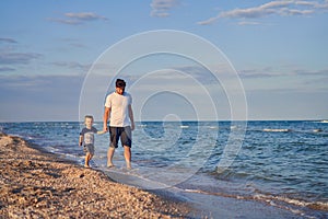 Young Caucasian dad with little son walk warm summer day along the sea coast. Summer family vacation concept. Friendship father