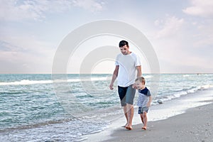 Young Caucasian dad with little son walk warm summer day along the sea coast. Summer family vacation concept. Friendship father