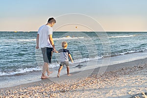 Young Caucasian dad with little son walk warm summer day along the sea coast. Summer family vacation concept. Friendship father