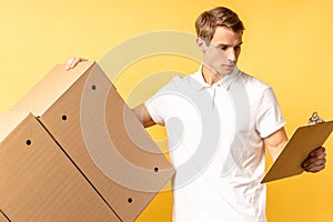 Young caucasian delivery man holding and carrying cardbox isolated on yellow background