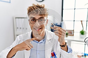 Young caucasian doctor man holding earing aid at the clinic smiling happy pointing with hand and finger