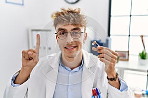 Young caucasian doctor man holding earing aid at the clinic smiling with an idea or question pointing finger with happy face,