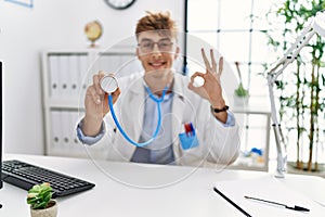 Young caucasian doctor man wearing doctor uniform and using stethoscope at the clinic doing ok sign with fingers, smiling friendly