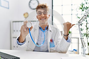 Young caucasian doctor man wearing doctor uniform and using stethoscope at the clinic pointing thumb up to the side smiling happy