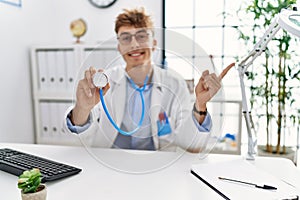 Young caucasian doctor man wearing doctor uniform and using stethoscope at the clinic smiling happy pointing with hand and finger