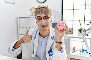 Young caucasian doctor man wearing doctor uniform holding piggy bank at the clinic smiling happy and positive, thumb up doing