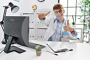 Young caucasian doctor man working at the clinic approving doing positive gesture with hand, thumbs up smiling and happy for