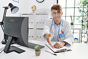 Young caucasian doctor man working at the clinic smiling friendly offering handshake as greeting and welcoming