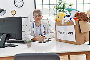 Young caucasian doctor man working at the clinic with toys donations box for children thinking attitude and sober expression