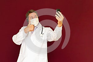 Young caucasian doctor with stethoscope, medical mask talking with patient online red background