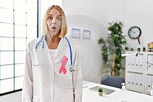 Young caucasian doctor woman wearing pink cancer ribbon scared and amazed with open mouth for surprise, disbelief face