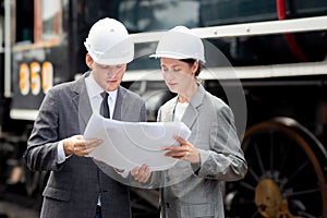 Young caucasian engineer man and woman in suit checking train looking blueprint in station.