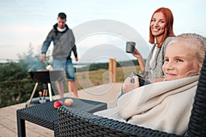 A young Caucasian family couple with a baby daughter sits on the terrace in chairs and drinks hot drinks. Against the