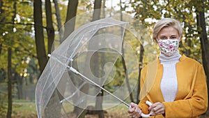 Young caucasian fashionable woman with face mask spinning transparent umbrela in the park in autumn.