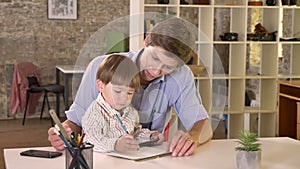 Young caucasian father holding his son and teaching him how to write, sitting in modern office