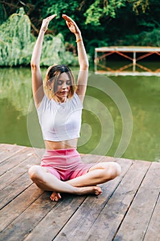 Young caucasian fitness woman doing yoga, meditate outdoors, on wooden pier lake on summer day