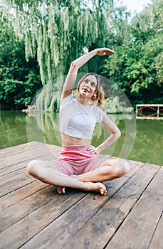 Young caucasian fitness woman doing yoga, meditate outdoors, on wooden pier lake on summer day