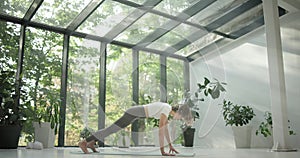 young caucasian fitness woman practicing yoga on a yoga mat in a modern studio with plants and large forest or garden