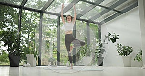 young caucasian fitness woman practicing yoga on a yoga mat in a modern studio with plants and large forest or garden