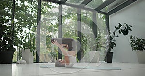 young caucasian fitness woman practicing yoga on a yoga mat in a modern studio with plants and large forest or garden