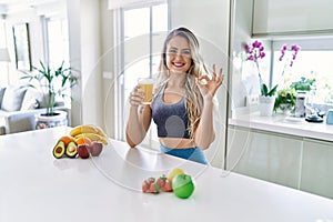 Young caucasian fitness woman wearing sportswear drinking healthy orange juice doing ok sign with fingers, smiling friendly