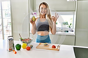 Young caucasian fitness woman wearing sportswear preparing healthy salad at the kitchen doing ok sign with fingers, smiling
