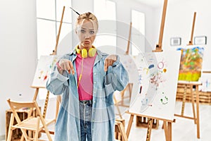 Young caucasian girl at art studio pointing down looking sad and upset, indicating direction with fingers, unhappy and depressed